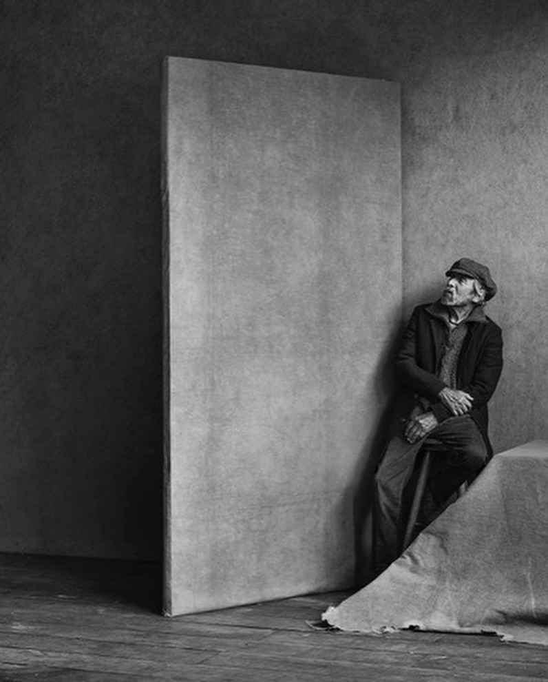 Monochrome portrait of an elderly man with a cap sitting in a corner, cane in hand, looking up, with light grey walls.
