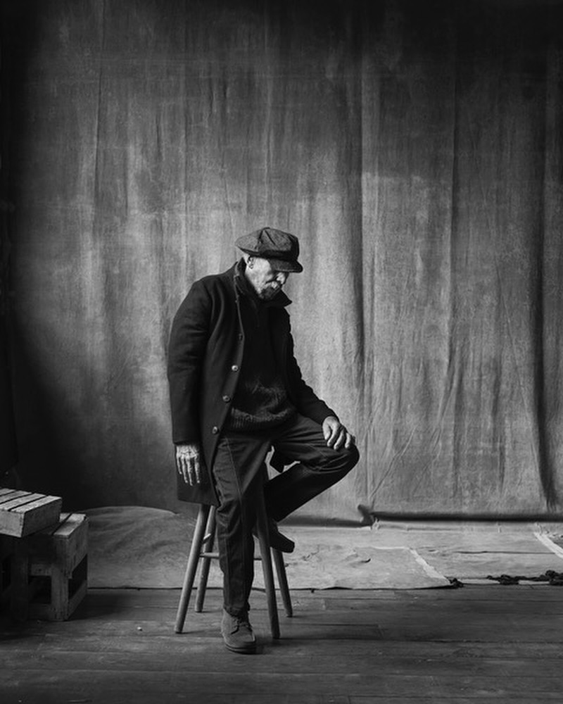 Monochrome portrait of a bearded adult male wearing a coat and cap, seated on a stool in a photo studio with a draped backdrop.