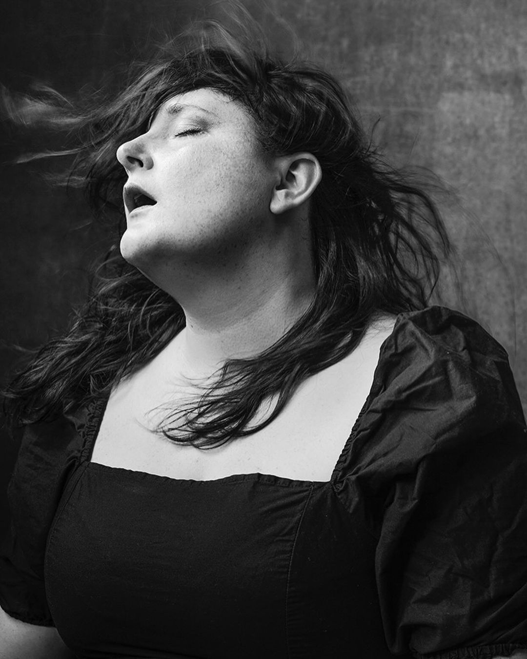 Black and white portrait of a woman with windswept dark hair and a square-necked dress, eyes closed, and mouth open in a dark studio.