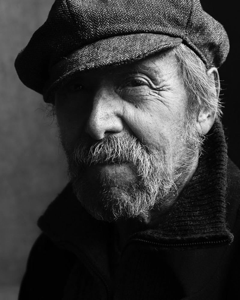 Black and white portrait of an adult male with a grey beard and mustache wearing a tweed cap, photographed in a dimly lit indoor setting.