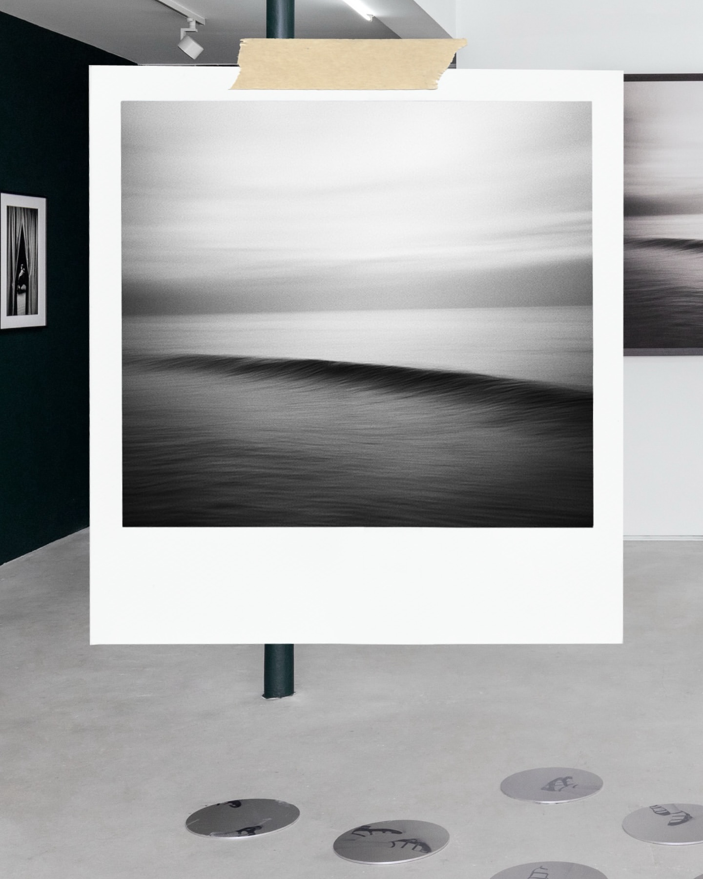 Monochrome seascape photograph on display at an art gallery, showcasing calm ocean waves and sky, taped to a dark support pole.