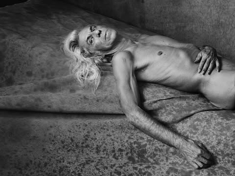 Monochrome portrait of an elderly adult male with long gray hair lying on a textured surface in a dimly lit indoor environment.