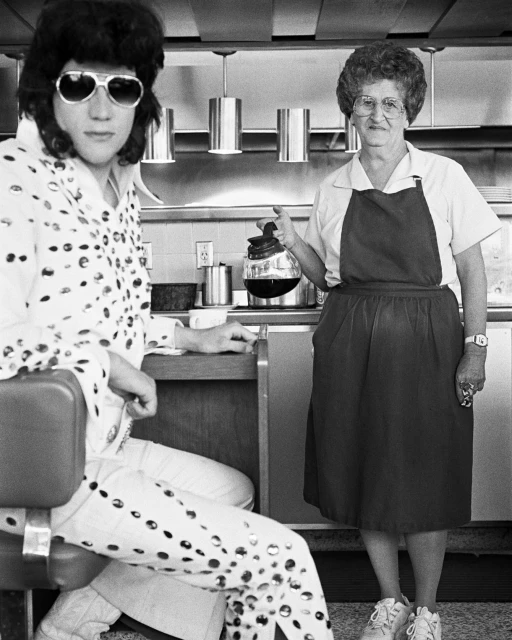 Black and white shot of a Elvis impersonator seated in a diner while a waitress pours him coffee, vintage aesthetic.