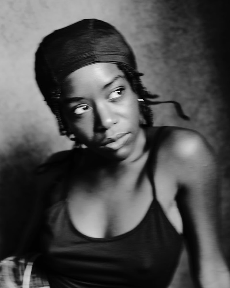 Monochrome portrait of a black woman wearing a black tank top and durag looking away with soft, diffused lighting in a studio setting.