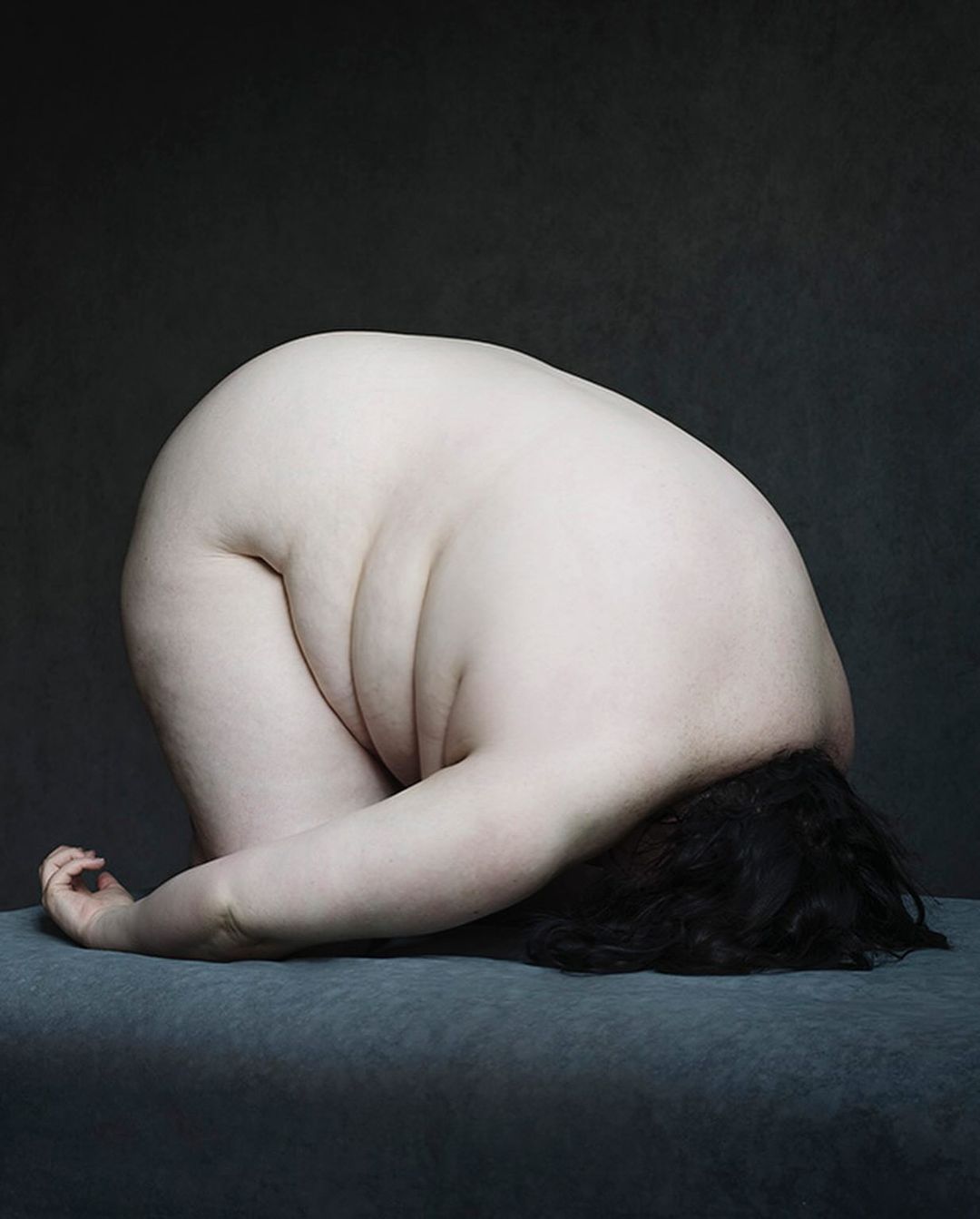 Overweight woman with dark hair in a head-down pose on a gray surface in a dimly lit studio setting, creating an intimate and introspective image.