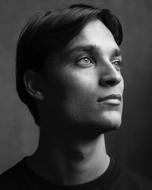 Black and white portrait of a young adult male with dark hair looking upward in a studio setting with soft lighting and dark background.