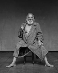 Elderly man with long white beard and hair sitting barefoot on a stool wearing a tweed coat