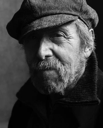 Close-up portrait of an elderly man with a beard wearing a flat cap and dark jacket captured in moody dramatic lighting by Stephan Vanfleteren