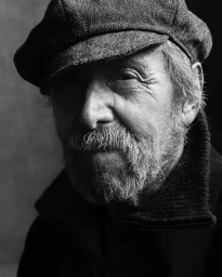 Close-up portrait of an elderly man with a beard wearing a flat cap and dark jacket captured in moody dramatic lighting by Stephan Vanfleteren