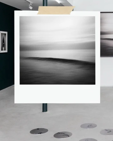 Black and white photograph of an ocean wave displayed as a polaroid print on a pole in an art gallery.