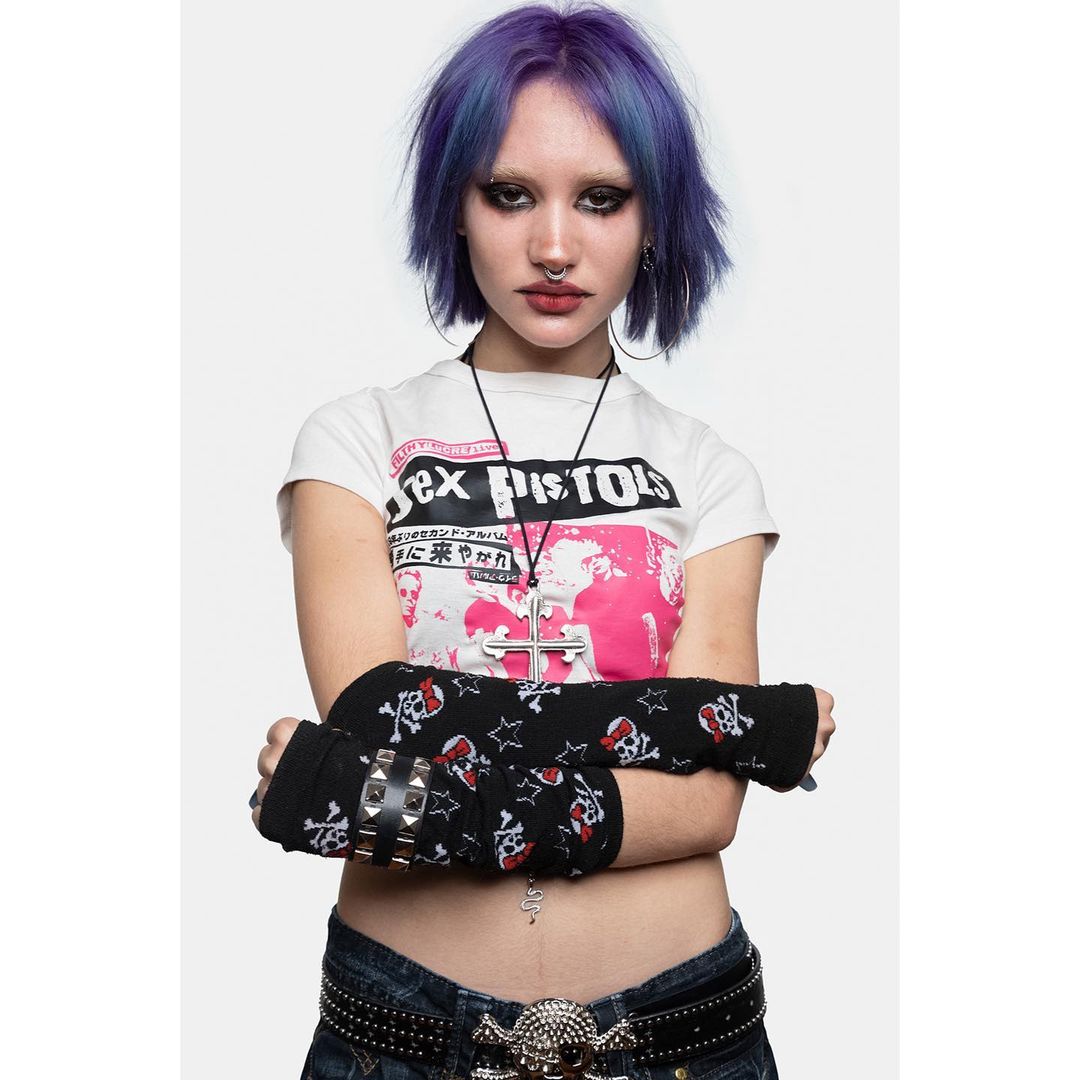 Young woman in a punk style wearing a graphic tee, black and red skull-printed arm warmers, and a belt with a skull buckle, against a white backdrop.