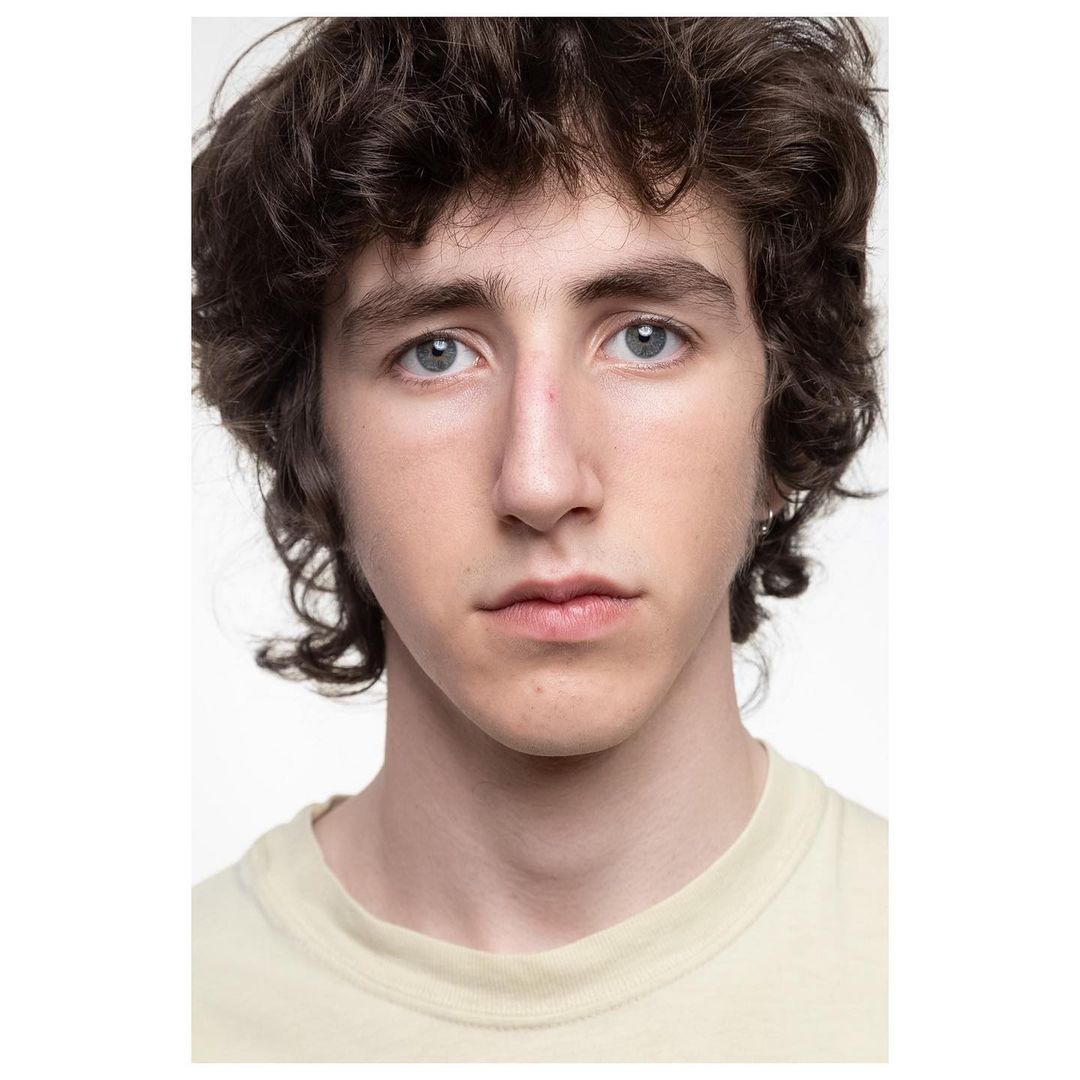 Close-up portrait of a young adult male with curly brown hair and light eyes, wearing a pale yellow shirt, against a white background.
