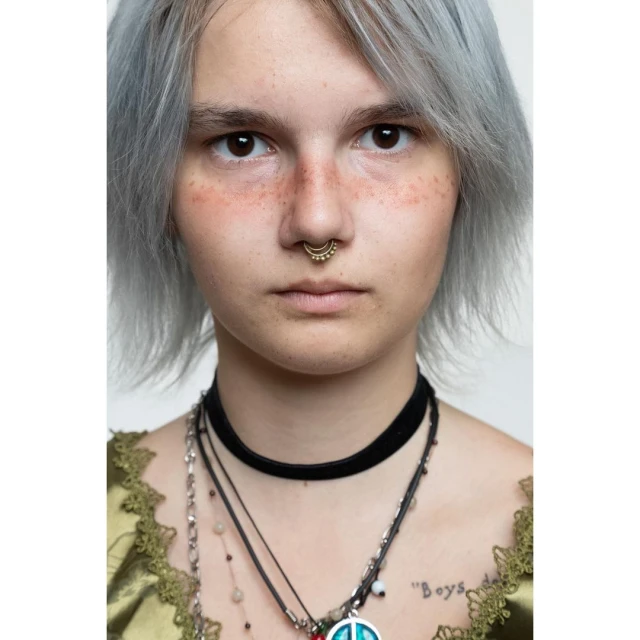 Close-up portrait of a young adult with light hair, brown eyes, a nose ring, wearing multiple necklaces, and a lace-trimmed top against a white backdrop.