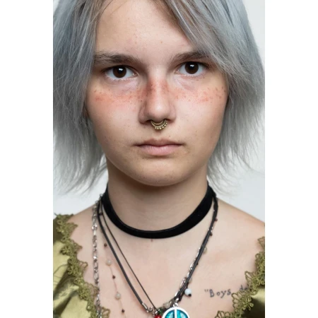 Close-up portrait of a young adult with light hair, brown eyes, a nose ring, wearing multiple necklaces, and a lace-trimmed top against a white backdrop.