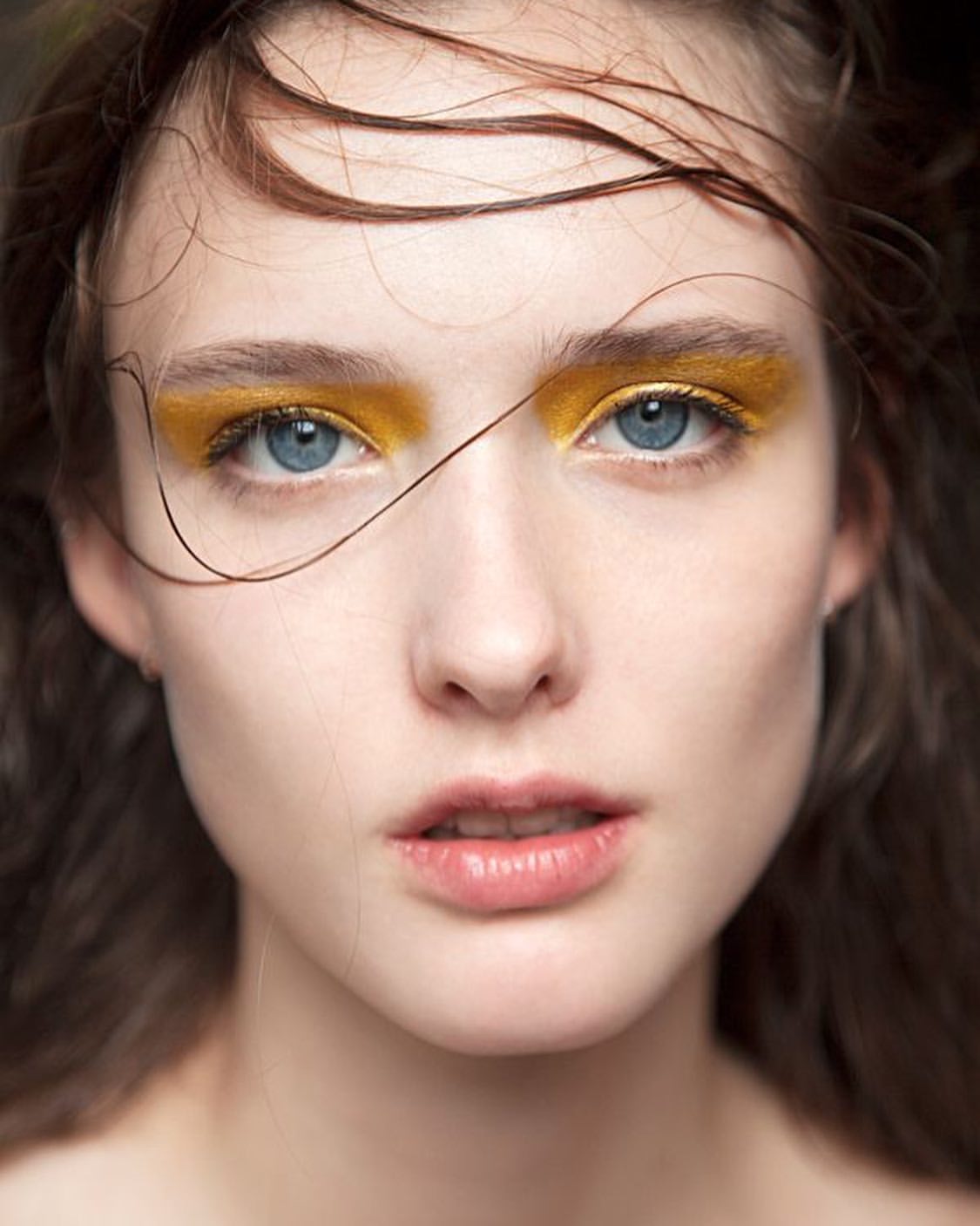 Close-up portrait of a woman with fair skin and blue eyes featuring bold yellow eyeshadow, styled wet hair, and a soft, diffused light.