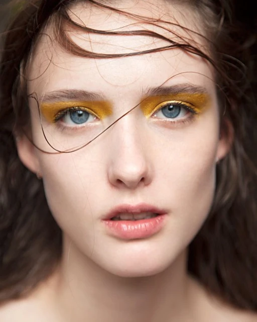 Close-up portrait of a woman with fair skin and blue eyes featuring bold yellow eyeshadow, styled wet hair, and a soft, diffused light.