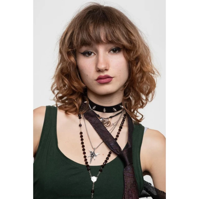 Portrait of a young adult woman with auburn curly hair, dark green top, and multiple necklaces against a white studio backdrop.