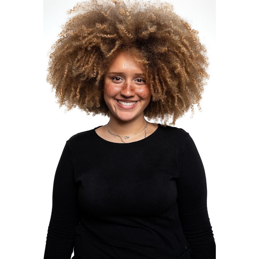 Portrait of a smiling woman with voluminous curly hair and freckles wearing a black long-sleeve top against a white background.