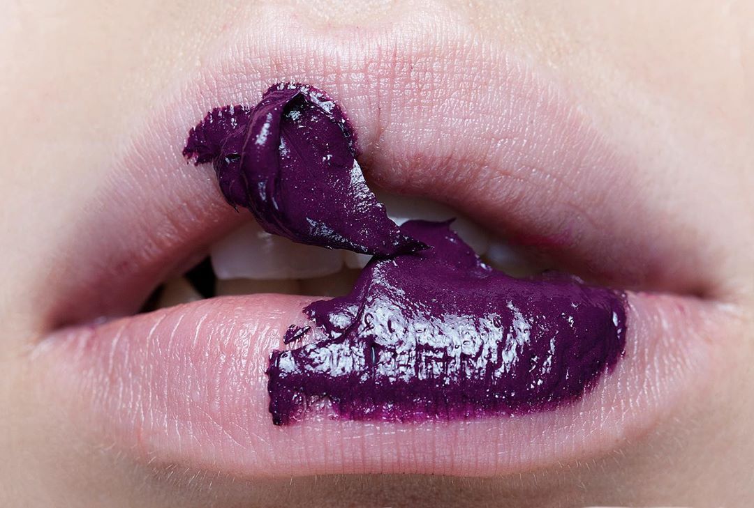 Close-up of a woman's lips with dark purple lip gloss smeared on the upper and lower lips, highlighting cosmetic beauty.