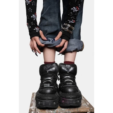 Standing woman wearing platform boots, blue jeans, and skull-print long-sleeve shirt, posing on a wooden surface against white background.