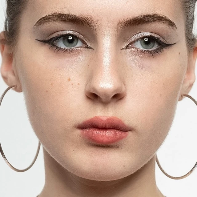 Close-up portrait of a young adult woman with light skin, gray eyes, dark eyeliner, and large hoop earrings against a white background.