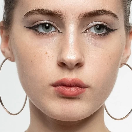 Close-up portrait of a young adult woman with light skin, gray eyes, dark eyeliner, and large hoop earrings against a white background.