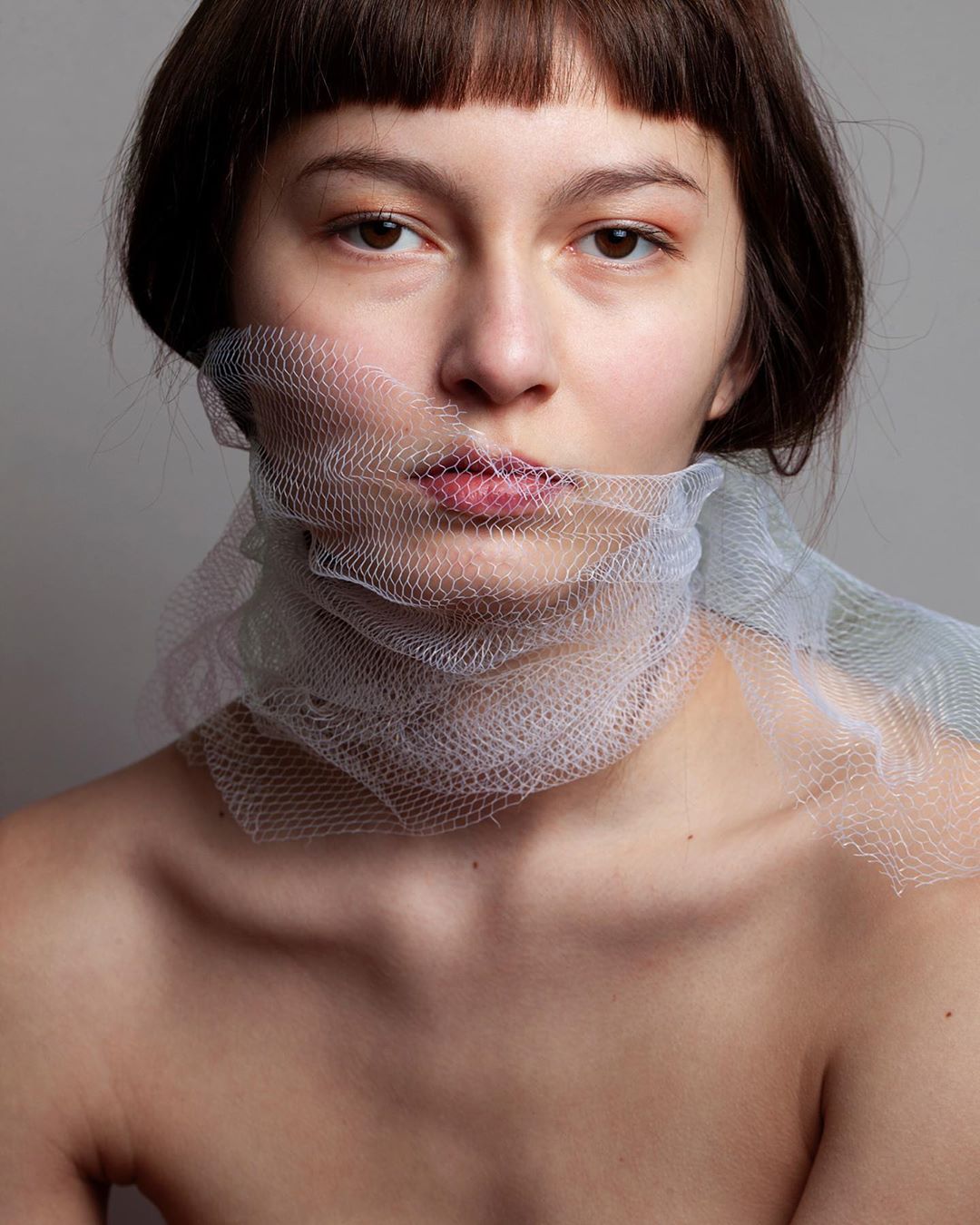 Portrait of a fair-skinned woman with brown hair and bangs, partially veiled by a textured, translucent white fabric around her face.