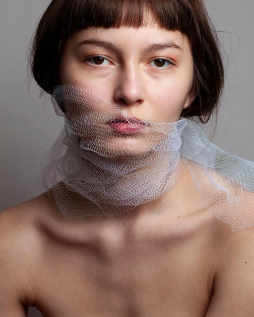 Portrait of a fair-skinned woman with brown hair and bangs, partially veiled by a textured, translucent white fabric around her face.
