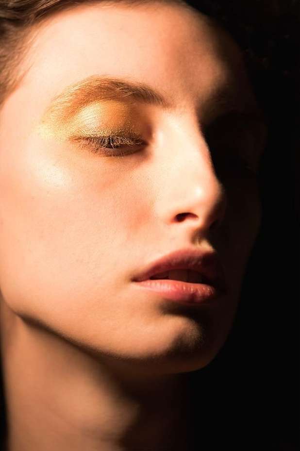 Close-up portrait of an adult female with closed eyes, warm skin tones, and gold eye shadow against a dark, contrasting background.