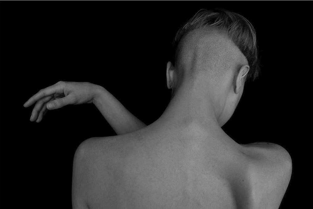 Monochrome portrait of a woman with an undercut hairstyle, seen from the back, showcasing her bare back against a black background.