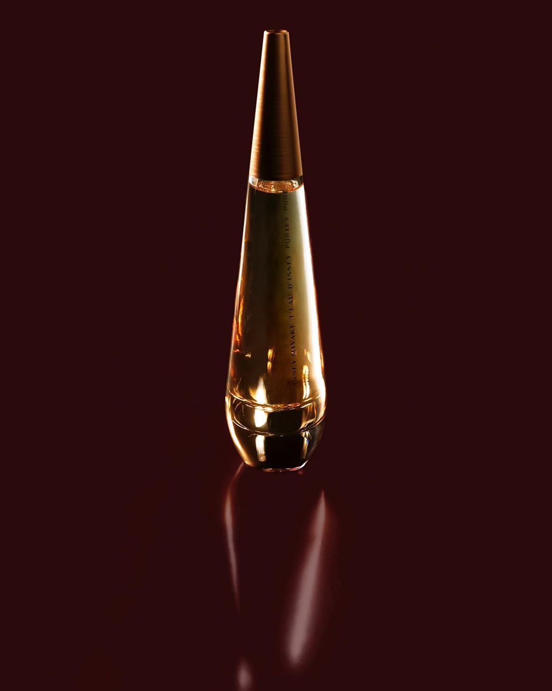 Elegant golden bottle of fragrance stands tall on a glossy maroon surface, accentuated by a gentle reflection.