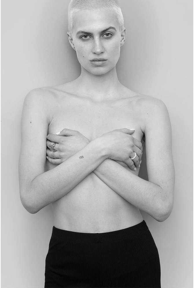 Portrait of a person with short blonde hair and rings, crossing arms against a plain background