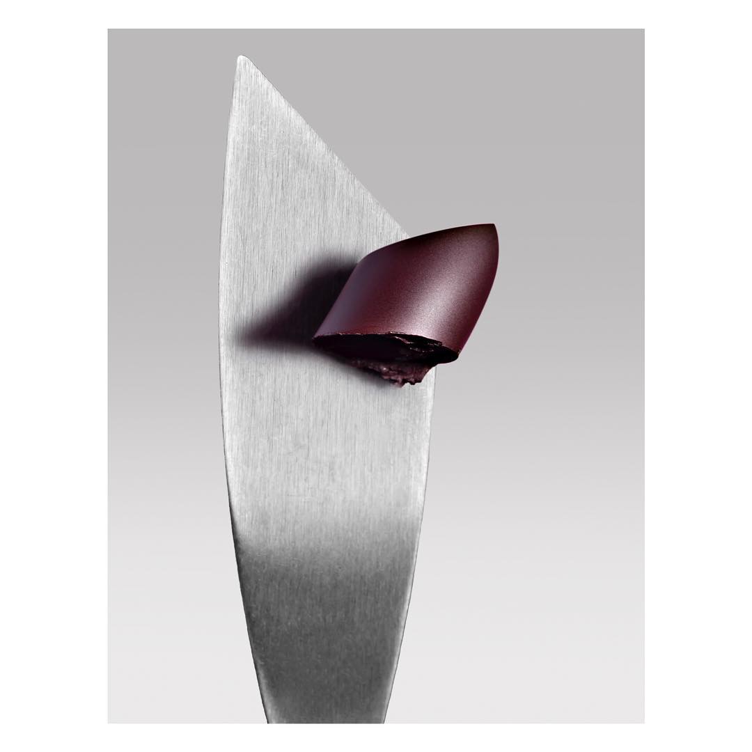 Close-up studio shot of deep red lipstick sample on a metal spatula against a grey backdrop, showcasing the cosmetic's texture and color.