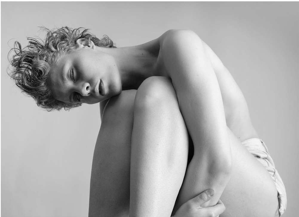 Monochrome portrait of a person with closed eyes, curly hair, hugging their knees in a photo studio setting against a neutral background.