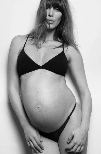 Black and white portrait of a pregnant adult female in underwear. Frontal pose. Indoor photo studio. Bangs hairstyle. Light background.