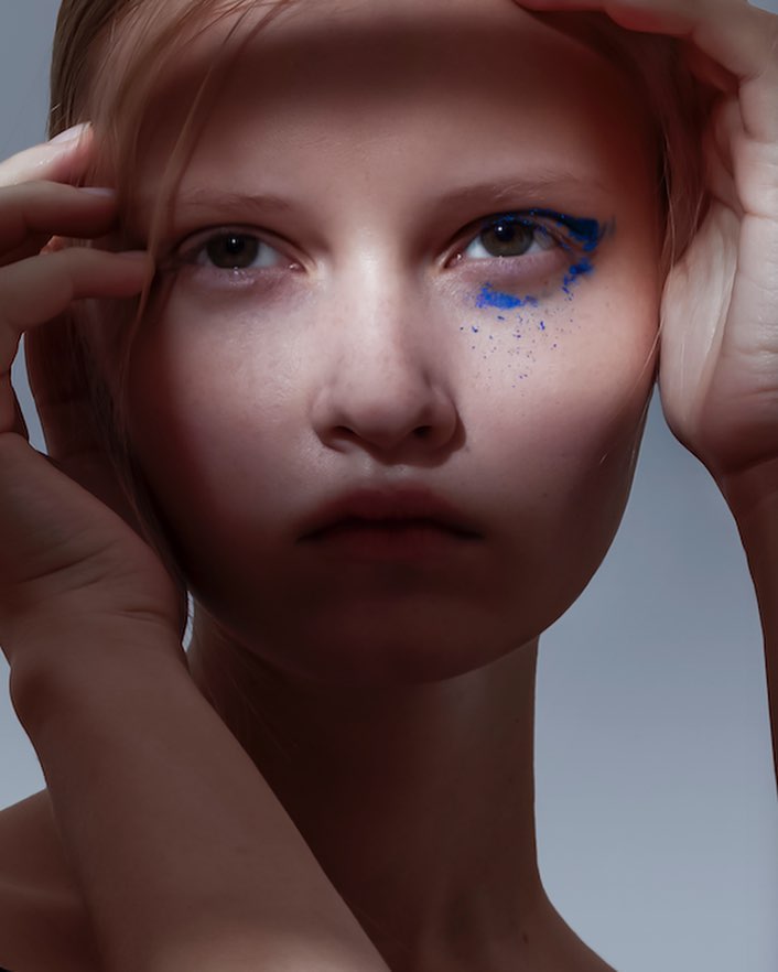Close-up of a young woman with blue makeup around her eye, fair skin, and light hair. Hands are visible near her face.