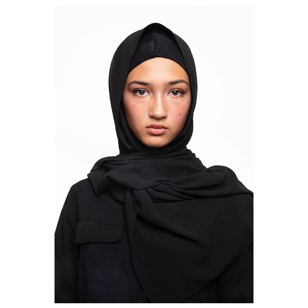 Headshot of a young woman with neutral expression wearing a black hijab and top against a bright white background in a studio setting.