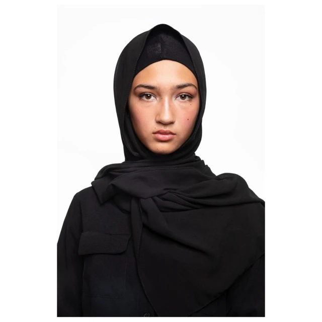 Headshot of a young woman with neutral expression wearing a black hijab and top against a bright white background in a studio setting.