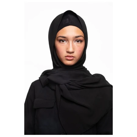 Headshot of a young woman with neutral expression wearing a black hijab and top against a bright white background in a studio setting.