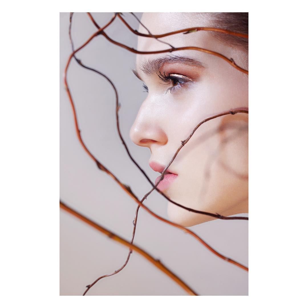 Close-up studio portrait of a young woman with soft makeup, framed by curving tree branches against a neutral background.