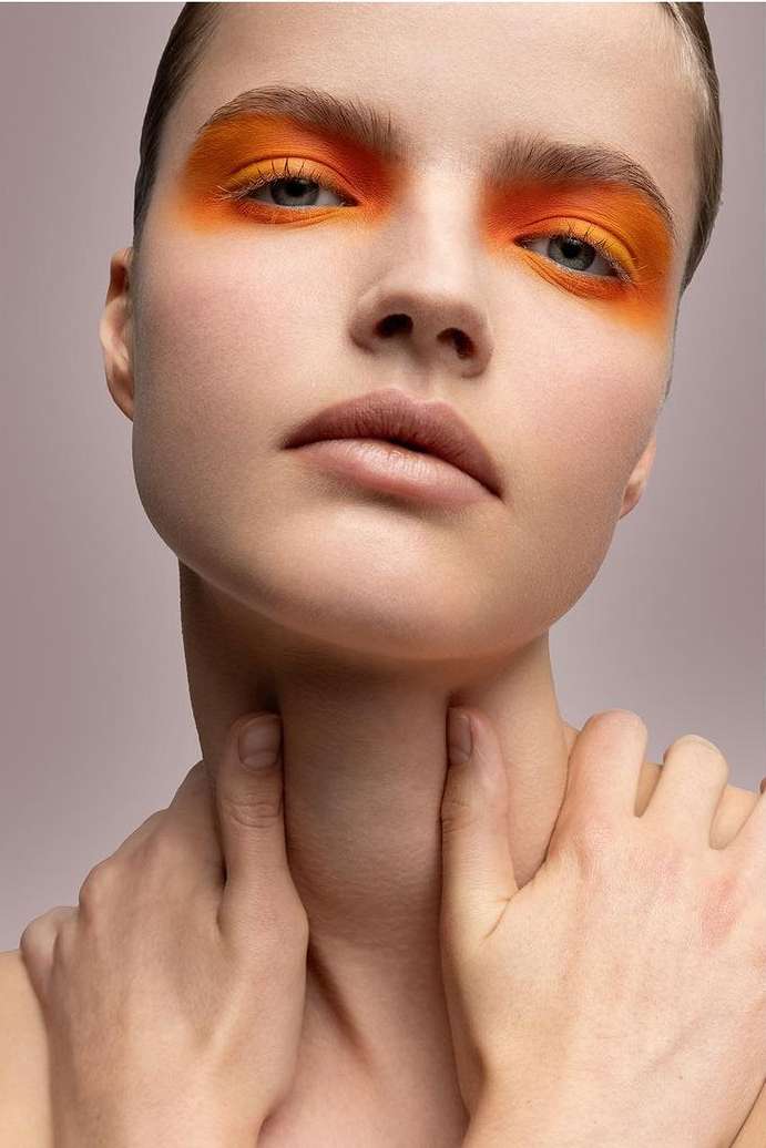 Close-up portrait of a young woman with orange eyeshadow, fair skin, and hands touching her neck against a pale pink background.