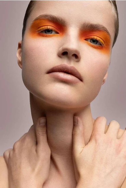 Close-up portrait of a young woman with orange eyeshadow, fair skin, and hands touching her neck against a pale pink background.