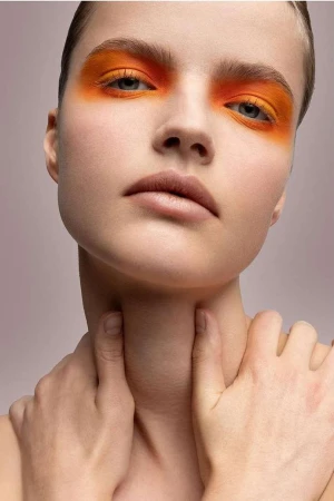 Close-up portrait of a young woman with orange eyeshadow, fair skin, and hands touching her neck against a pale pink background.