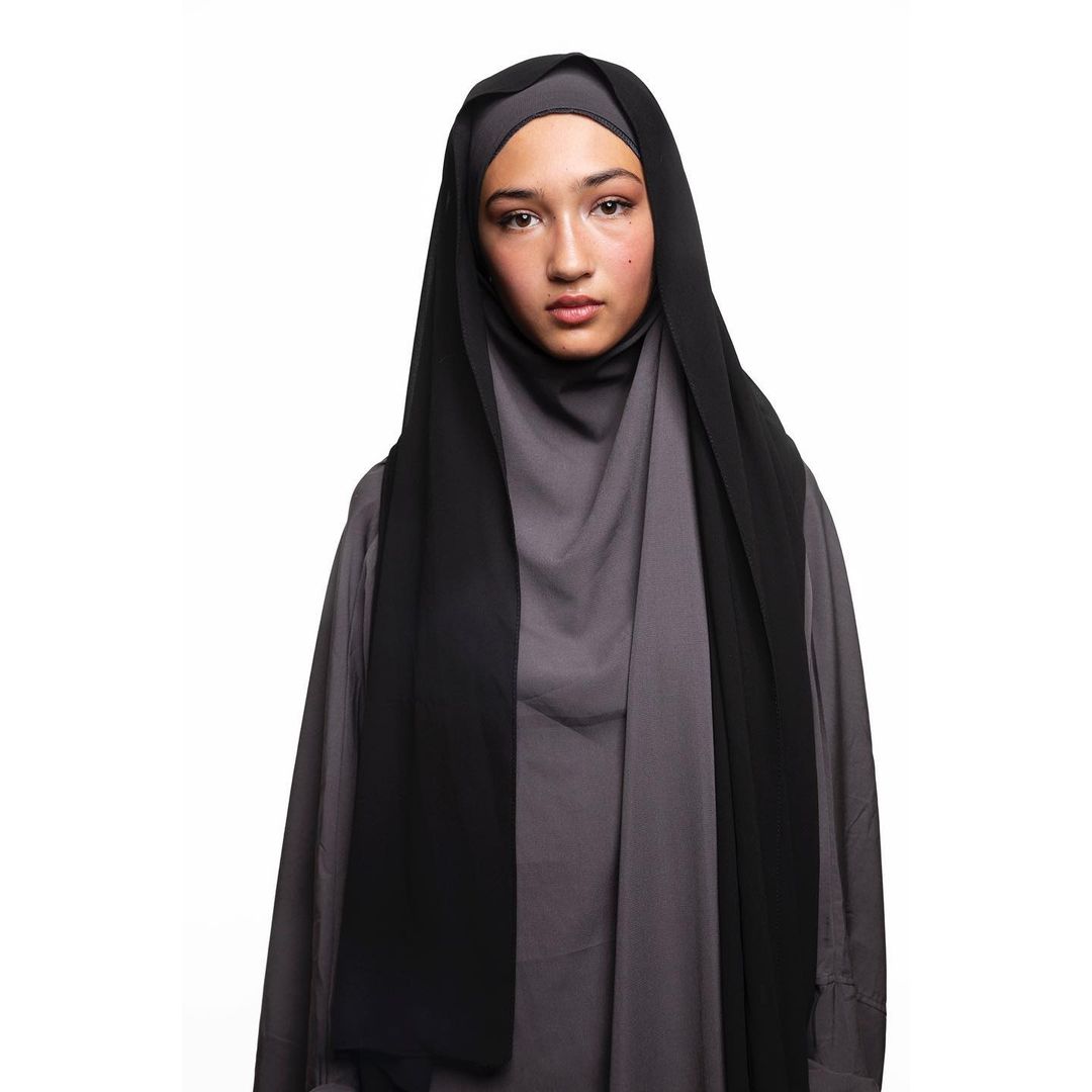 Portrait of a young woman wearing a black headscarf and gray tunic against a white backdrop in a fashion or beauty context.
