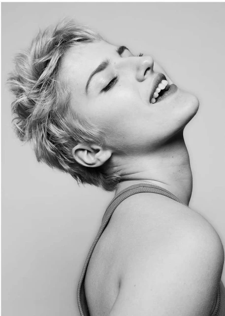 Black and white portrait of a young woman with short blonde hair, eyes closed, and mouth open in laughter, tilting her head back against a plain background.