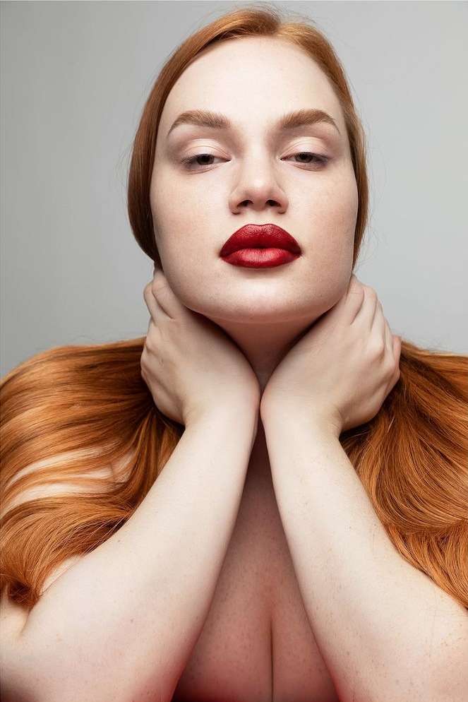 Headshot of a redhead woman with hands on her neck and dark red lipstick, posing against a neutral grey backdrop in a studio setting.