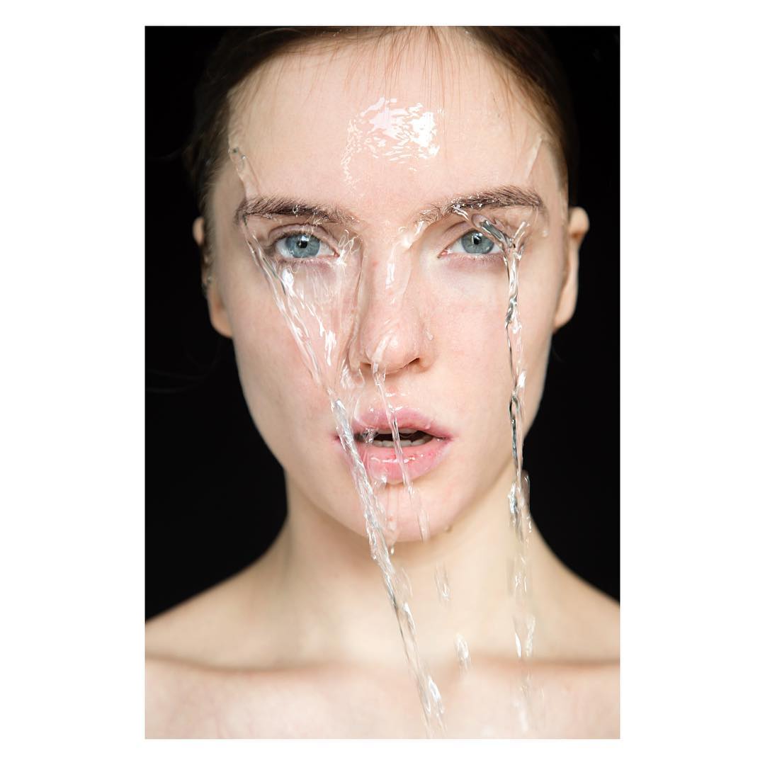 Close-up studio portrait of a young fair-skinned woman with light blue eyes, covered in glistening water with a stark black background.