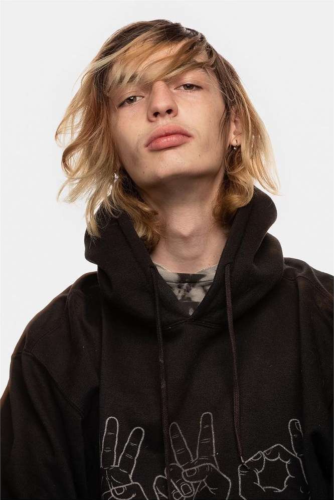 Headshot of a young adult with highlighted hair wearing a black hooded sweatshirt with hand gesture graphic on an isolated white background.