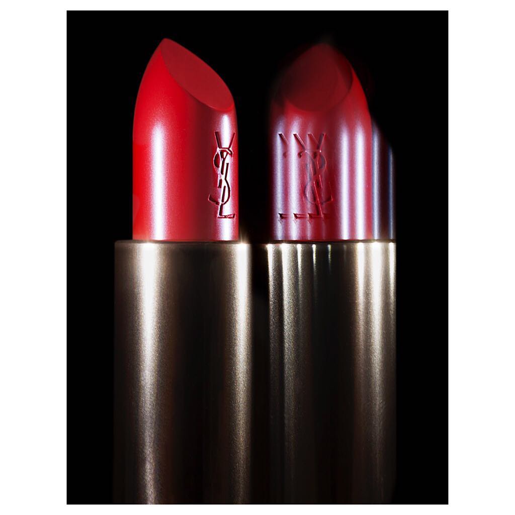 Two red Yves Saint Laurent lipsticks with gold tubes are prominently displayed against a black background in a beauty studio shot.
