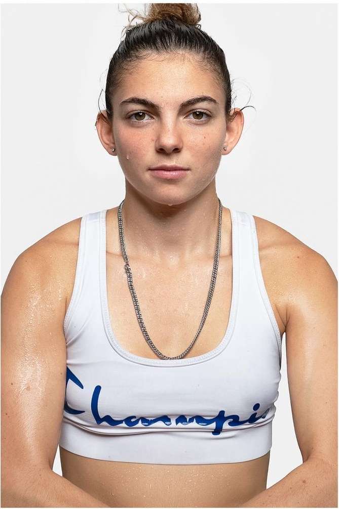 Portrait of a sweaty woman wearing a white sports bra and silver necklace against a white backdrop in an eye-level studio shot.